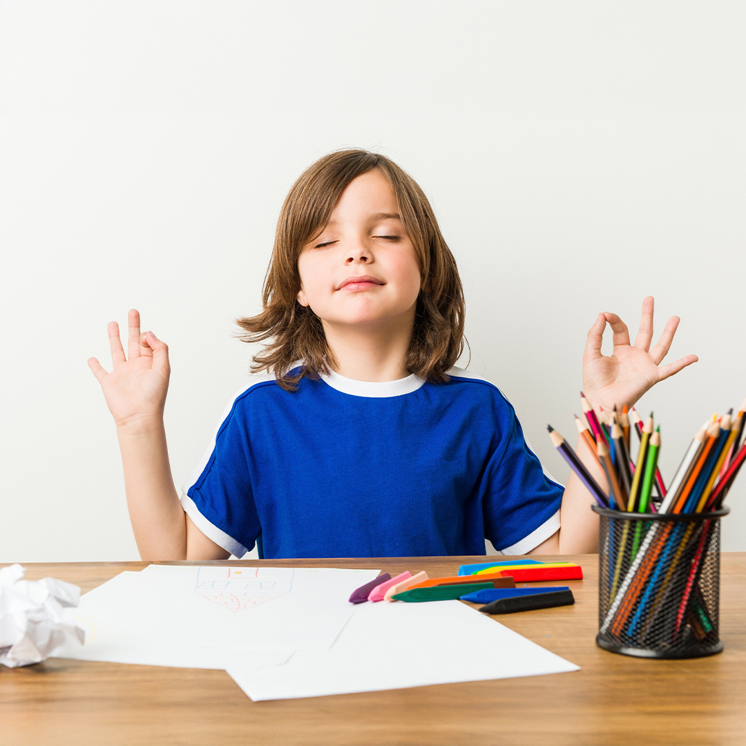 Kind sitzt mit geschlossenen Augen am Tisch und meditiert, daneben liegen bunte Stifte und Papier 20251215_DGK_Artikel-03_7Mind-Mindfulness-Classroom.jpg