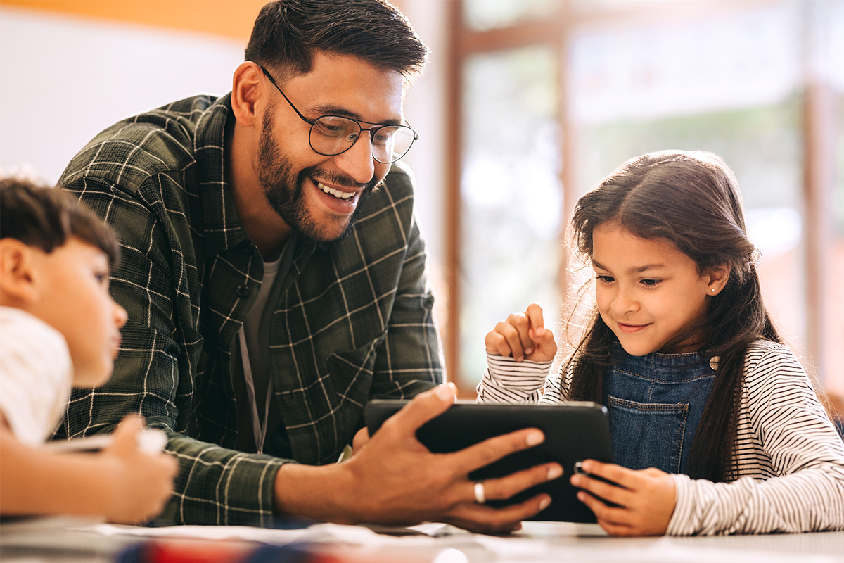 Ein erwachsener Mann mit Brille und kariertem Hemd schaut gemeinsam mit zwei Kindern auf ein Tablet. Die Szene spielt in einem hellen Klassenzimmer oder Schulgebäude und zeigt einen gemeinsamen Lernmoment mit digitalen Medien.