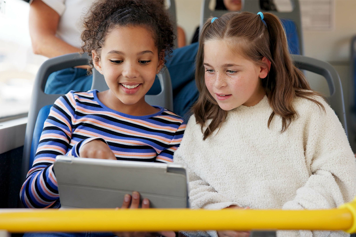Zwei Mädchen sitzen nebeneinander in einem Bus und schauen gemeinsam auf ein Tablet.