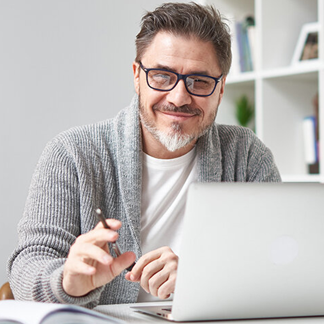 Mann mit Brille und grauer Strickjacke sitzt vor Laptop und blickt lächelnd in die Kamera DGK_Lehrer_AZwischen-Fake-News-und-wissenschaftlicher-Erkenntnis-header.jpg