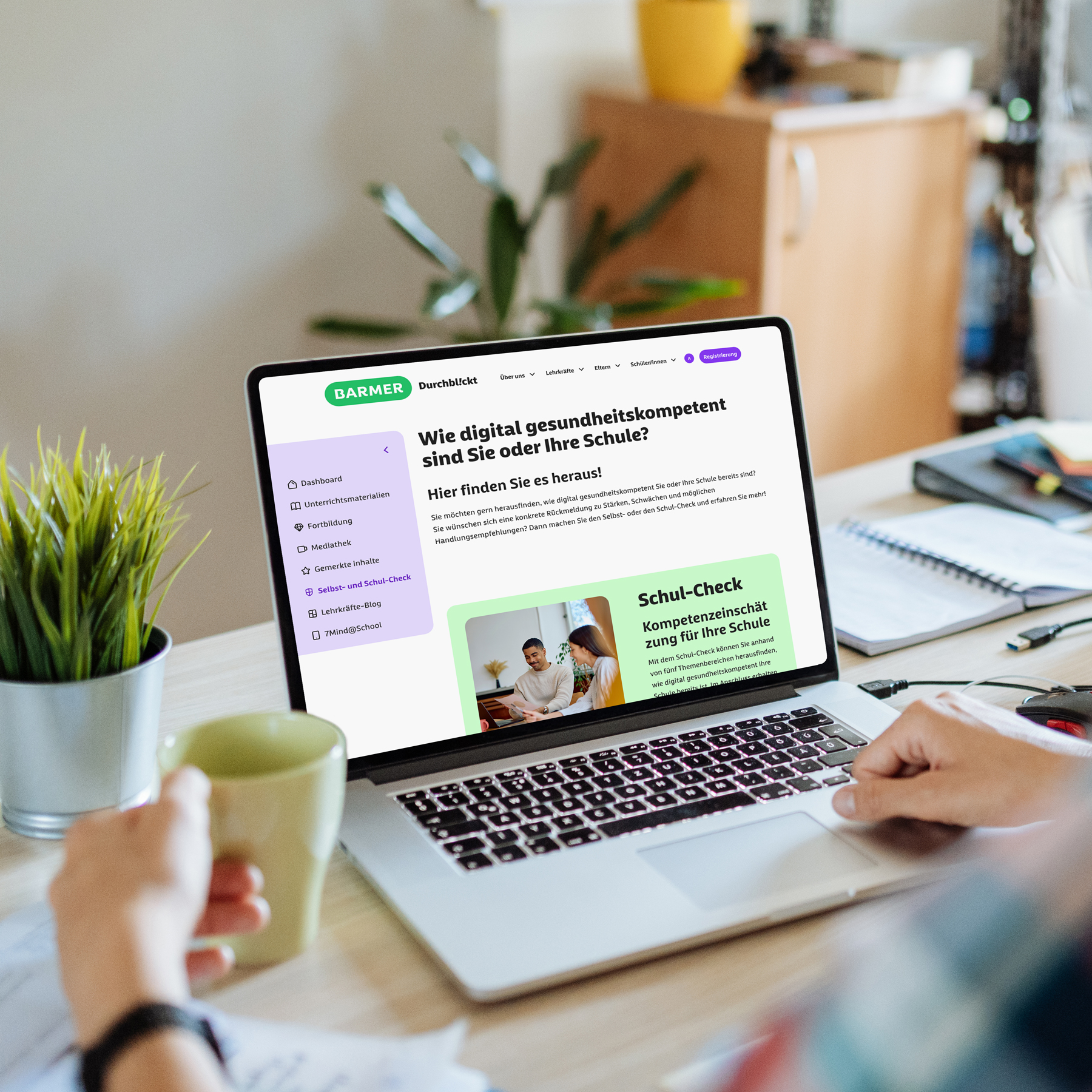 Lehrerin am Holzschreibtisch mit Laptop zeigt BARMER Durchblickt Schul-Check zur digitalen Gesundheitskompetenz, grüne Buttons, moderne Schulumgebung