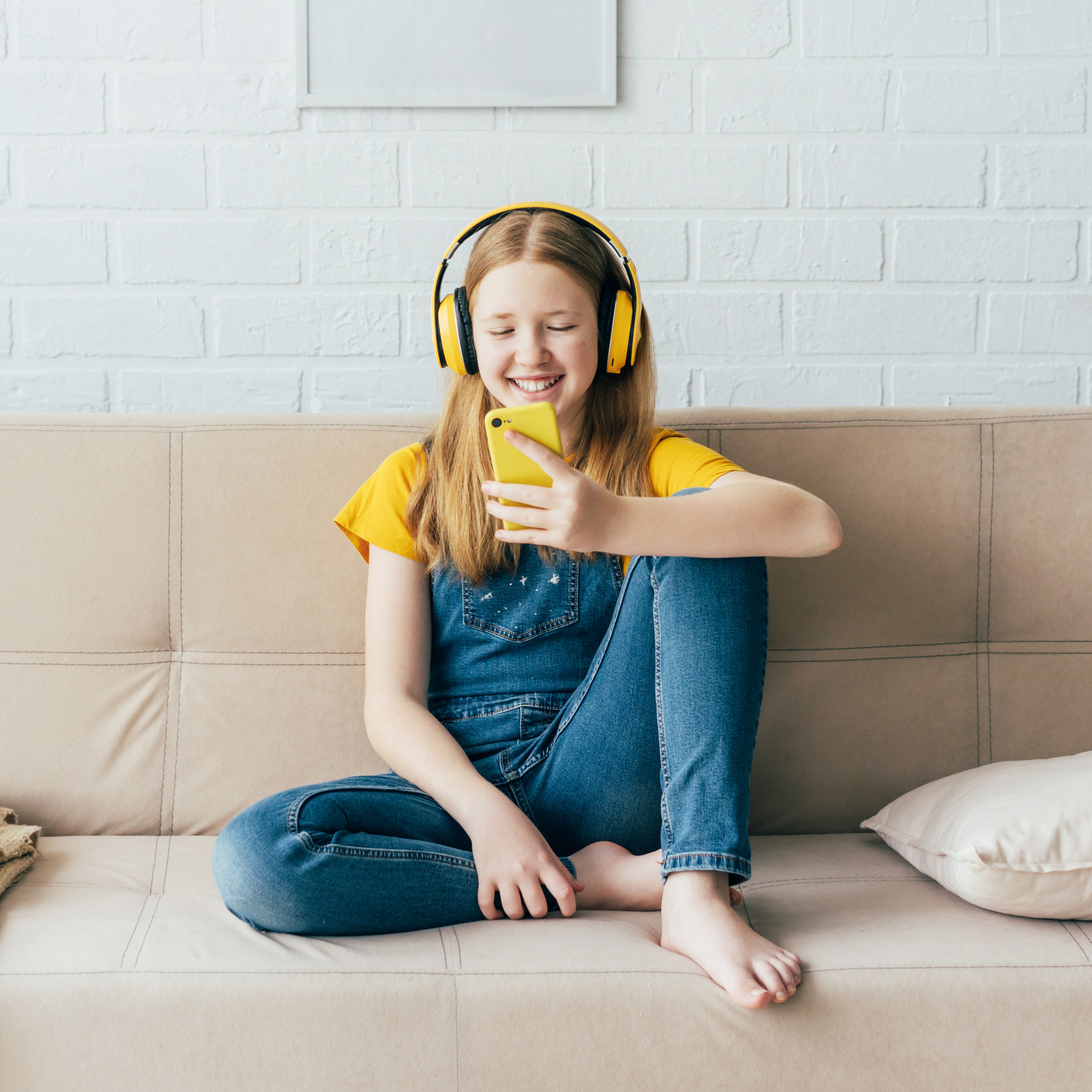 Schülerin hört Podcast mit gelben Kopfhörern und Smartphone auf beigem Sofa.