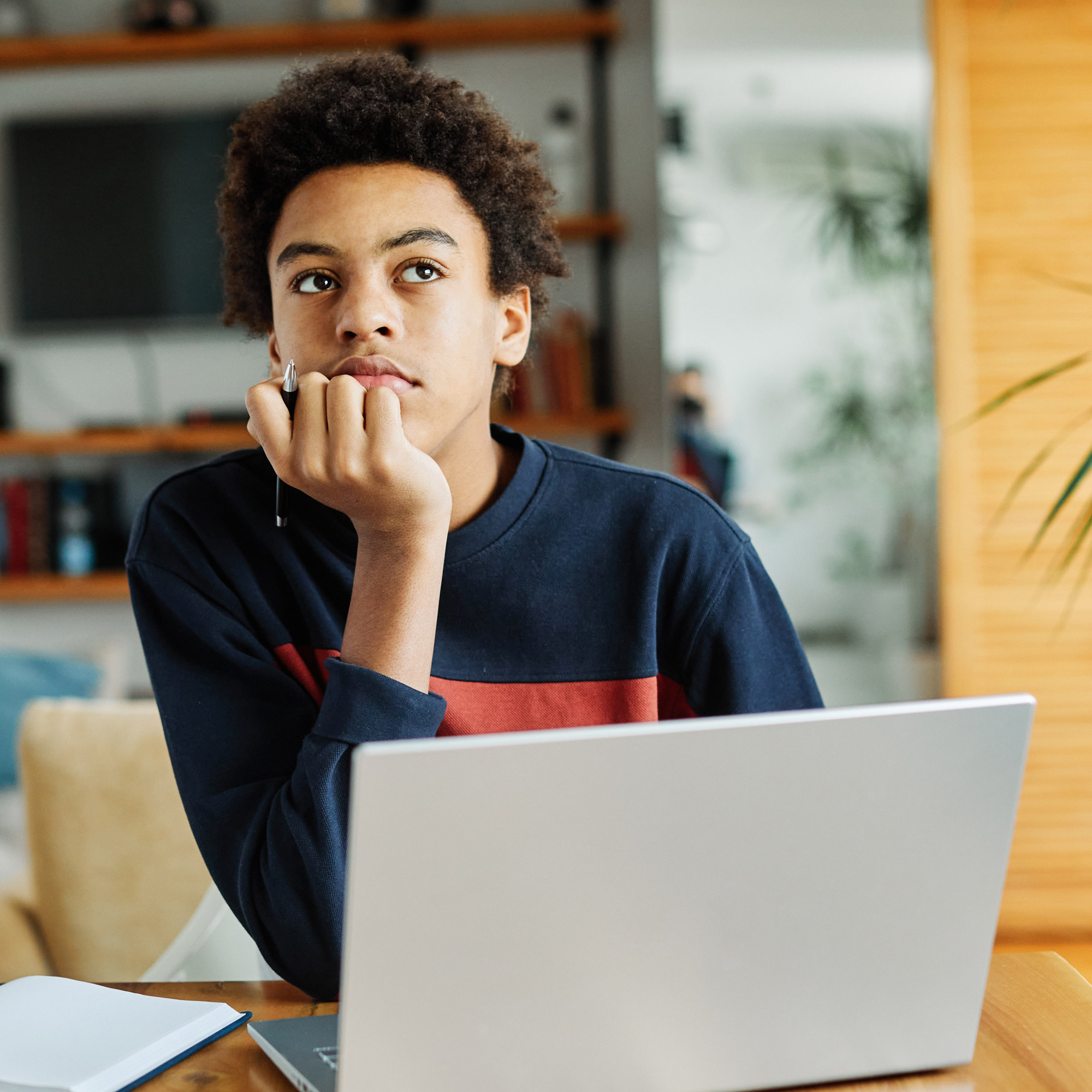Jugendliche Person mit Afro-Frisur nachdenklich am Laptop, Hand am Kinn, Notizbuch und Stift, konzentriertes Lernen