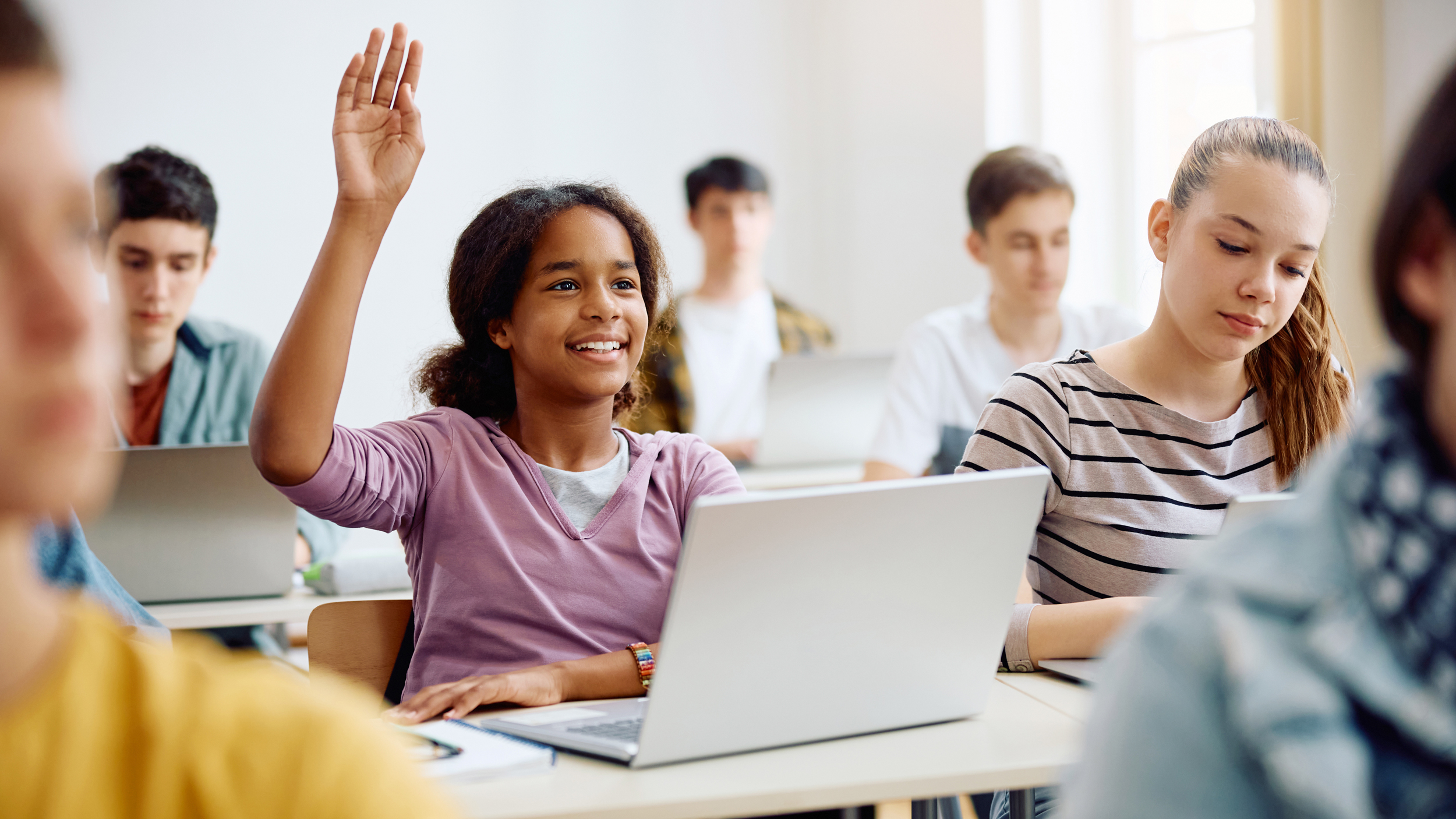 Schülerin mit Regenbogenarmband hebt Hand am Laptop-Schreibtisch, lächelt engagiert