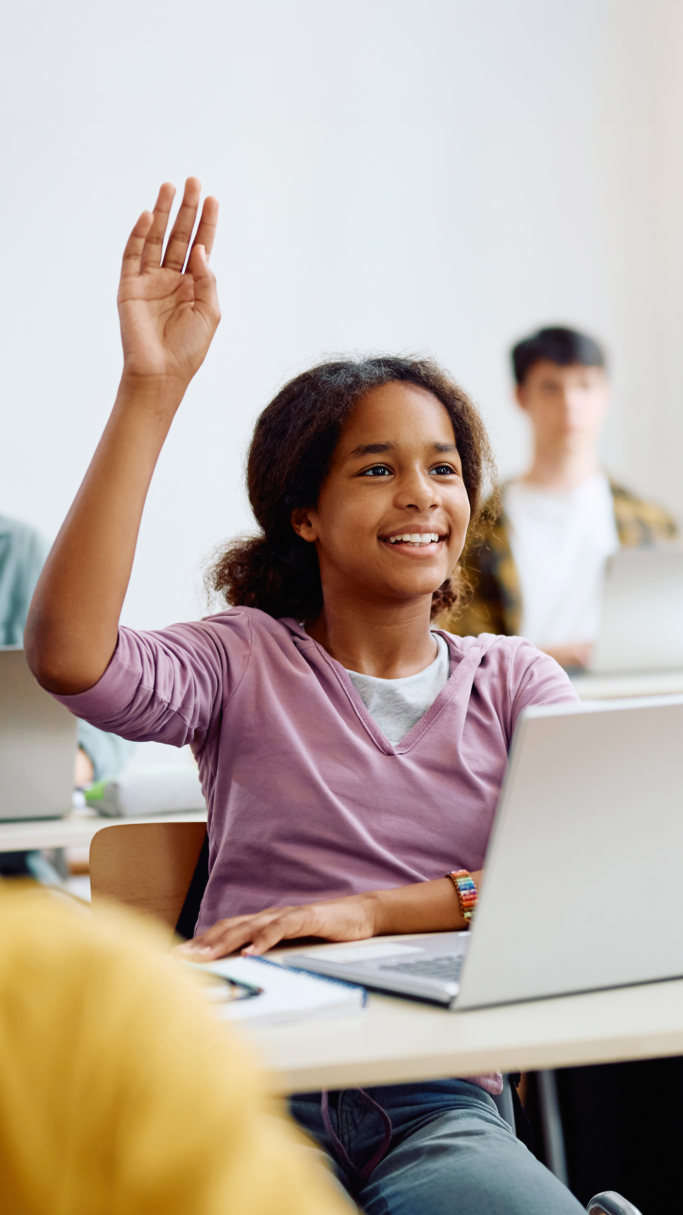 Schülerin mit dunklen Haaren hebt Hand im Klassenzimmer, andere Schüler arbeiten am Laptop